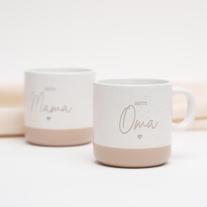 May include: Two white ceramic mugs with a speckled finish. The mug on the left says "Beste Mama" with a small heart. The mug on the right says "Beste Oma" with a small heart. Both mugs have a light brown band around the bottom.