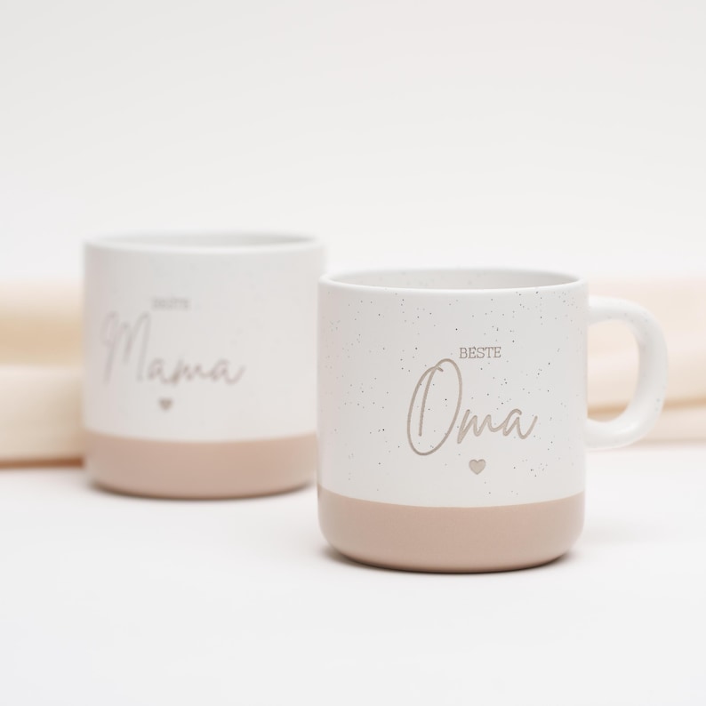 May include: Two white ceramic mugs with a speckled finish. The mug on the left says "Beste Mama" with a small heart. The mug on the right says "Beste Oma" with a small heart. Both mugs have a light brown band around the bottom.