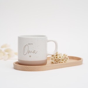 May include: White ceramic mug with a speckled finish and a brown bottom. The mug has the text "Beste Oma" with a small heart symbol.