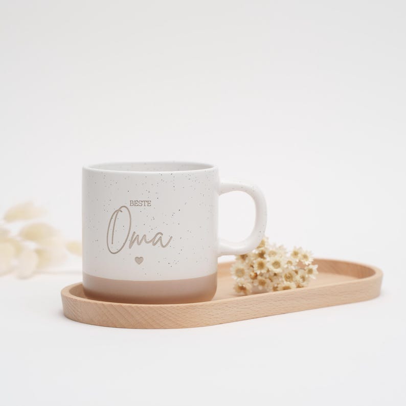 May include: White ceramic mug with a speckled finish and a brown bottom. The mug has the text "Beste Oma" with a small heart symbol.