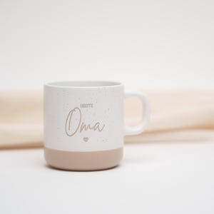 May include: White ceramic mug with a speckled finish and a pink base. The mug features the text "Beste Oma" with a small heart symbol.