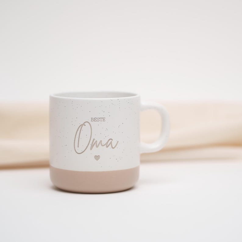 May include: White ceramic mug with a speckled finish and a pink base. The mug features the text "Beste Oma" with a small heart symbol.
