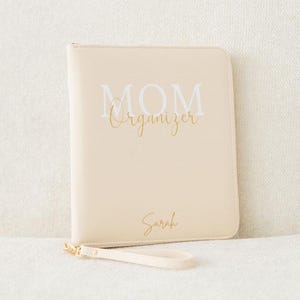 Könnte beinhalten: Ein beiger Lederorganizer mit der Aufschrift "MOM Organizer" in Weiß und dem Namen "Sarah" in goldener Kursivschrift.
