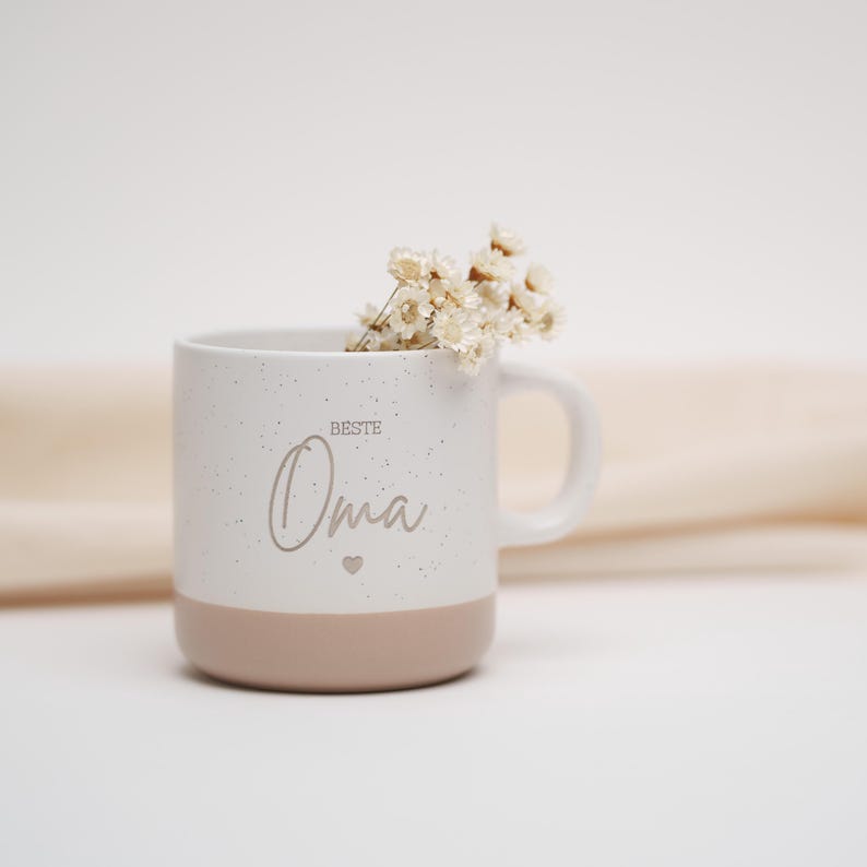 May include: White ceramic mug with a speckled finish and a light pink base. The mug features the text "Beste Oma" in a cursive font with a small heart symbol.