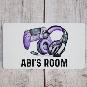 May include: A white sign with purple and black gaming accessories, a video game controller and headphones, with the text "ABI'S ROOM" in black.
