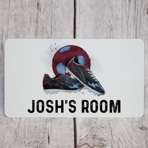 May include: A white rectangular sign with rounded corners features a soccer ball and cleats illustration. The ball is maroon and blue, and the cleats are black and blue. The text "JOSH'S ROOM" is printed in bold black letters.