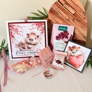 Könnte beinhalten: Ein Geschenkset mit einer Grußkarte mit einem Cartoon-Igel in einer Hängematte, Tee, Schokolade und einem kleinen Dekorationsartikel. Die Karte trägt den Text "Kleine Auszeit". Das Set enthält ein rosa Kneipp-Produkt und eine Lindt Schokoladenwaffel.