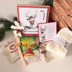 Könnte beinhalten: Ein Geschenkset bestehend aus einer quadratischen Schachtel mit Rentier-Illustration und dem Text Mach mal Pause, einer Karte mit Rentier, einer Packung Van Houten Kakao, einem Holzlöffel, einem Badesalzbeutel, zwei Schokoriegeln und weißen Kuschelsocken.