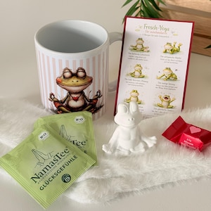Könnte beinhalten: Eine weiße Keramiktasse mit rosa vertikalen Streifen und einem Cartoon-Frosch, der meditiert. Das Bild enthält auch zwei Teebeutel, eine weiße Froschfigur, ein rotes Bonbon und eine Karte mit dem Titel "Frosch-Yoga".