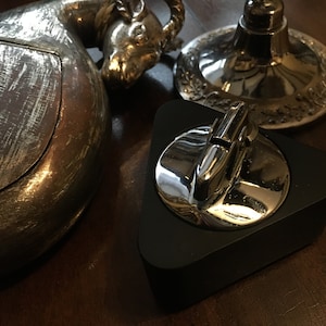 May include: A silver-coloured metal ashtray shaped like a ram's head, a silver-coloured metal base with a decorative design, and a black triangular lighter stand with a chrome lighter.