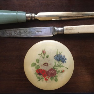 May include: A set of two silver letter openers with bone and jade handles, resting on a round, white alabaster trinket box with a floral design.