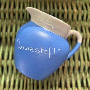 May include: A blue ceramic jug with a white rim and handle, inscribed with the word "Lowestoft" in white script. The jug is set against a green woven background.