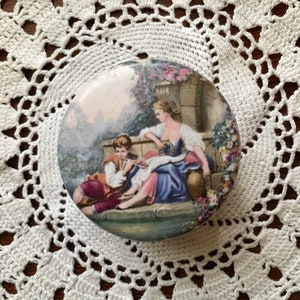May include: A round porcelain trinket box featuring a painted scene of a couple in a garden setting. The woman is wearing a blue dress and the man is wearing a brown and red outfit. The box has a white background with a floral design around the edge.