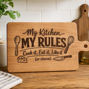 Puede incluir: Tabla de cortar de madera con el texto "My Kitchen My Rules" e ilustraciones temáticas de cocina. La tabla presenta una cuchara, un batidor, queso y un cuchillo. La tabla tiene un asa y está hecha de madera clara.
