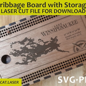 Op de afbeelding: Een houten cribbage-bord met opslag, met een lasergesneden ontwerp van Lake Winnipesaukee, New Hampshire. Het bord heeft een kompas en coördinaten voor het meer. De tekst "SVG.PDF" en "@ASHCAT.LASER" zijn ook zichtbaar.