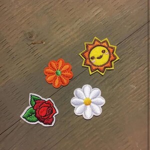 Può includere: Quattro toppe ricamate: un fiore arancione con centro verde, un sole giallo sorridente, una rosa rossa con foglie verdi e una margherita bianca con centro giallo. Le toppe sono su una superficie di legno.