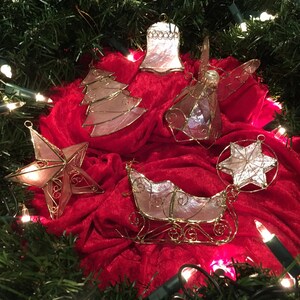 May include: A selection of Christmas decorations, including a star, bell, angel, tree, sleigh, and star-shaped ornament, on a red velvet surface. The ornaments have a gold wire frame and a pearlescent finish. Green garland and fairy lights surround the decorations.