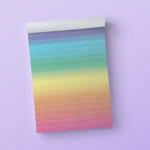 Op de afbeelding: Een regenboogkleurig notitieboekje met gelinieerde pagina's. Het notitieboekje is bovenaan opgerold en ligt op een lichtpaarse achtergrond.