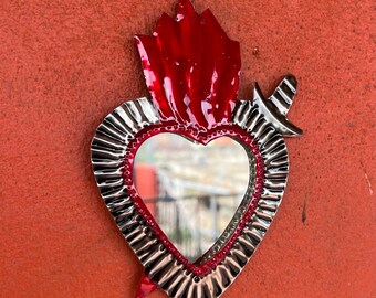 May include: A heart-shaped mirror with a red flame design at the top and a silver-colored metal frame. A silver-colored sword pierces the heart. The mirror is hanging on a red wall.