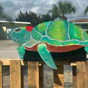 May include: A large, hand-painted wooden turtle sculpture with a green and teal shell, red accents, and a brown underside. The sculpture is mounted on a wooden fence, with a sunny sky and palm trees in the background. The turtle is a decorative garden art piece.