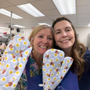 May include: Two oven mitts with a light blue background and a pattern of fried eggs and faces. The mitts are quilted and appear to be made of fabric. The mitts are being held up by two people.