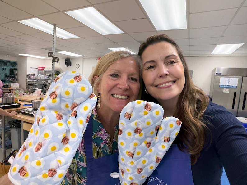 May include: Two oven mitts with a light blue background and a pattern of fried eggs and faces. The mitts are quilted and appear to be made of fabric. The mitts are being held up by two people.