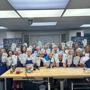 May include: A group of people in a kitchen holding up oven mitts. The mitts are white with a floral pattern. The people are wearing aprons and smiling. A wooden table is in front of them.