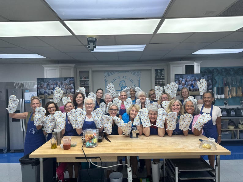May include: A group of people in a kitchen holding up oven mitts. The mitts are white with a floral pattern. The people are wearing aprons and smiling. A wooden table is in front of them.