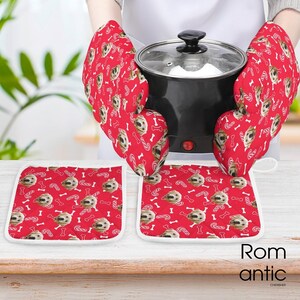 May include: Red oven mitts and pot holders with a repeating pattern of dog faces, bones, and candy canes. The mitts are holding a black pot with a glass lid. The pot holders are square with white trim. The word "Romantic" is in the bottom right corner.