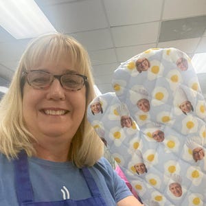 May include: Light blue oven mitt with a pattern of fried eggs and faces. The mitt is quilted and has a loop for hanging. The faces are of people wearing chef hats. The background is a light blue color.