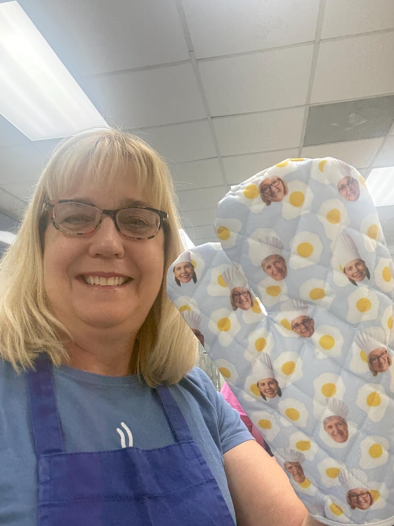 May include: Light blue oven mitt with a pattern of fried eggs and faces. The mitt is quilted and has a loop for hanging. The faces are of people wearing chef hats. The background is a light blue color.