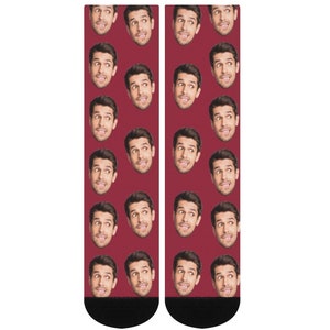 K&ouml;nnte beinhalten: Ein Paar rote Socken mit einem sich wiederholenden Muster eines M&auml;nnergesichts. Die Socken haben schwarze B&uuml;ndchen.