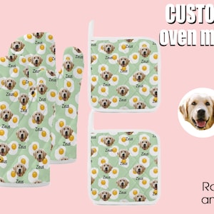 Op de afbeelding: Een set van drie groene ovenwantjes met een witte rand. De wantjes hebben een herhalend patroon van gele eieren en een hondenkop met de naam "Zeus" op elk ei. De wantjes zijn ontworpen voor gebruik in de keuken.