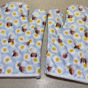 May include: A pair of quilted oven mitts with a light blue background. The mitts are patterned with yellow fried eggs and faces of chefs wearing white hats. The edges are white with a white loop for hanging.