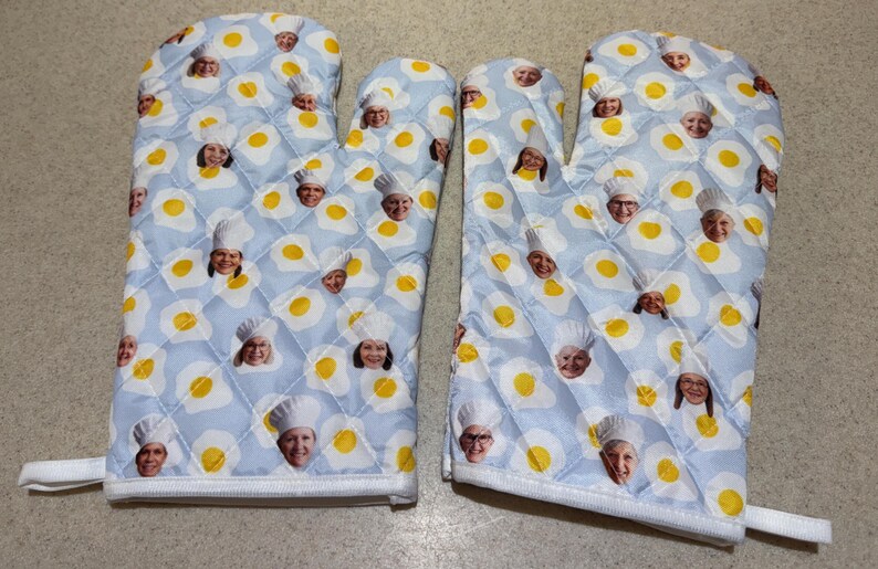 May include: A pair of quilted oven mitts with a light blue background. The mitts are patterned with yellow fried eggs and faces of chefs wearing white hats. The edges are white with a white loop for hanging.