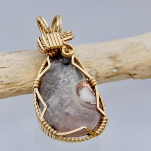May include: A teardrop-shaped pendant with a gold-colored wire wrap. The pendant features a gray and pink stone with white and copper accents. The pendant is displayed on a piece of driftwood against a light gray background.