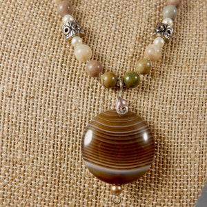 May include: A handcrafted necklace featuring a large, round, brown agate pendant with white and brown banding. The necklace includes a beaded strand of neutral-toned gemstones, pearls, and silver-toned metal accents. The pendant hangs from a silver-toned wire.