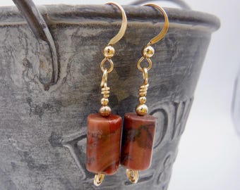 May include: A pair of dangle earrings featuring cylindrical red and brown jasper beads suspended from gold-toned metal hooks. Each earring includes a small gold-toned metal bead spacer above the stone cylinder and a simple wire loop at the base.