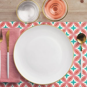 May include: A white dinner plate with a gold rim sits on a pink and teal patterned placemat. A white napkin with a gold fork and knife is to the left of the plate. A gold spoon is to the right of the plate. There is a glass of water and a glass of pink wine above the placemat.