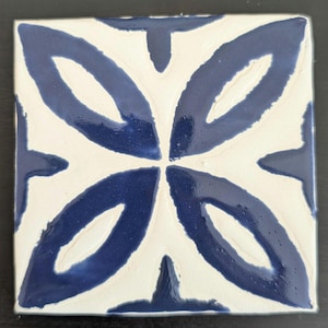 May include: A white ceramic tile with a blue floral design. The design features four stylized flower shapes with rounded edges.
