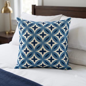 May include: A square throw pillow featuring a repeating geometric pattern of interlocking circles and four-pointed stars. The design uses a color palette of cream, light blue, and dark navy blue, creating a symmetrical and balanced visual effect.