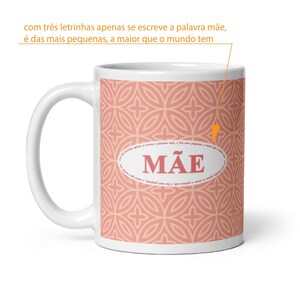 May include: A white ceramic mug featuring a repeating coral and light pink geometric pattern. Centered on the mug is an oval frame containing the word MÃE in large pink letters, surrounded by small Portuguese text.