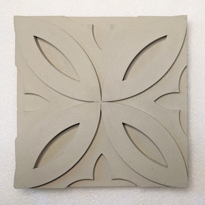 May include: A square, light gray tile with a raised floral design. The design features four large, stylized leaf shapes radiating from the center, with smaller, curved elements in the corners. The tile has a textured surface and a slightly rough edge.