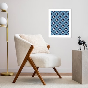 May include: A framed art print featuring a repeating pattern of blue and white floral shapes. The artwork is displayed on a white wall above a white armchair with wooden legs. A gold floor lamp and a black dog statue are also visible.