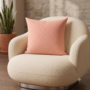 May include: A square throw pillow featuring a coral-colored fabric with a repeating white geometric pattern of intersecting circles and four-pointed stars. The pillow has a smooth texture and is displayed centered on a cream-colored armchair.