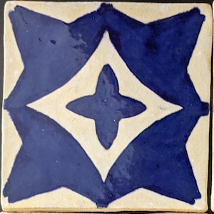 May include: A white ceramic tile with a blue geometric pattern. The pattern features a four-pointed star in the center, surrounded by four diamond shapes.