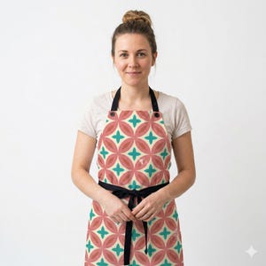 May include: An apron featuring a repeating geometric pattern of interlocking pink oval shapes and teal four-pointed stars on a cream background. The apron includes black neck and waist ties, providing a secure fit for various tasks.