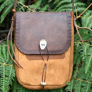 May include: A brown leather crossbody bag with a suede body and a decorative flap. The bag features a bone clasp and intricate stitching. The bag has a long strap for shoulder or crossbody wear. The bag is suitable for everyday use.