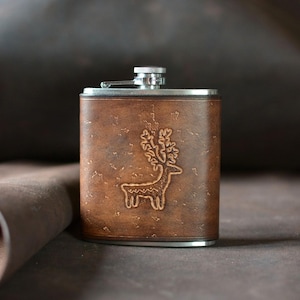 May include: A rectangular stainless steel flask wrapped in brown leather with a textured surface. The leather features an embossed graphic of a deer with large antlers centred on the front. The flask has a silver metal screw-top cap.