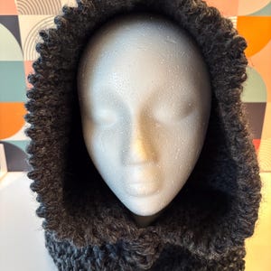 May include: A dark gray knitted cowl and hood combination. The hood is designed with a textured, ruffled edge, and the cowl section is a thick, close knit. The item is displayed on a mannequin head, showcasing its shape and fit.